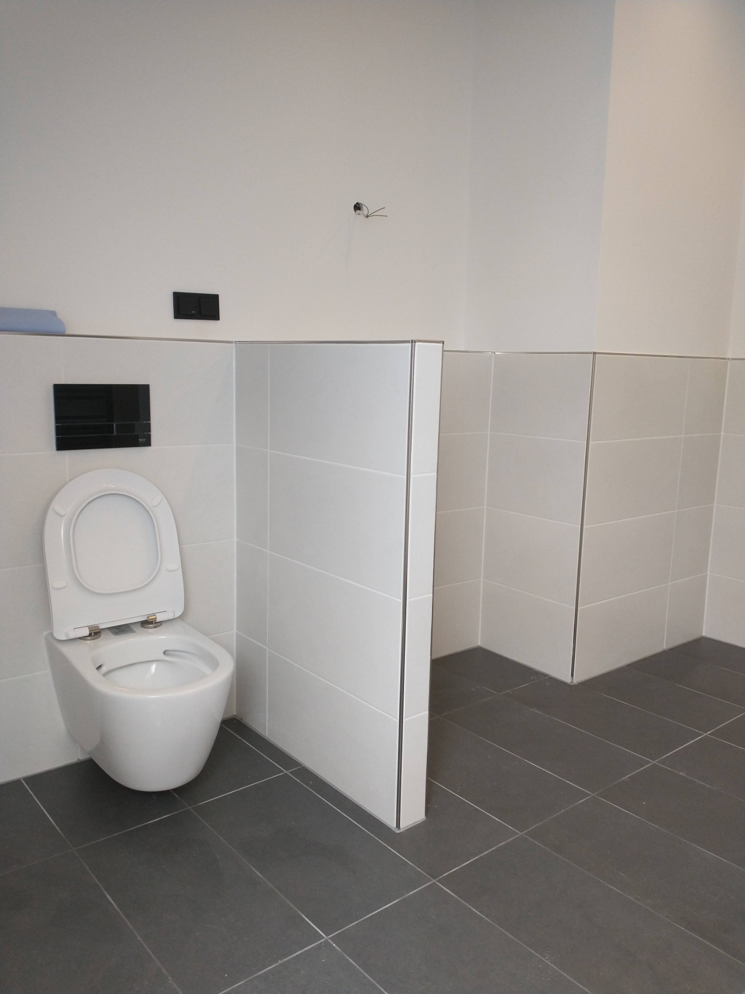 Nowoczesna toaleta podwieszana z otwartą deską w minimalistycznej łazience z białymi płytkami ściennymi i grafitową podłogą, widoczne czarne panele sterujące i otwór na kabel.