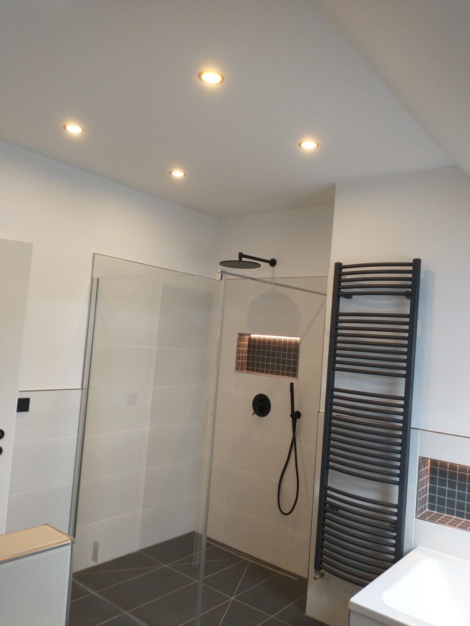 Nowoczesna łazienka z kabiną prysznicową typu walk-in, czarną armaturą, grzejnikiem drabinkowym i punktowym oświetleniem sufitowym.