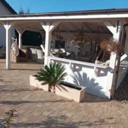 Biała altana z drewnianą konstrukcją, wyposażona w meble rattanowe i wiszące fotele, otoczona kostką brukową i roślinami w donicach w słoneczny dzień.