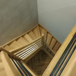 Widok z góry na drewniane schody z jasnego drewna z czarnymi, metalowymi poręczami, układające się w zygzak na tle szarej ściany.