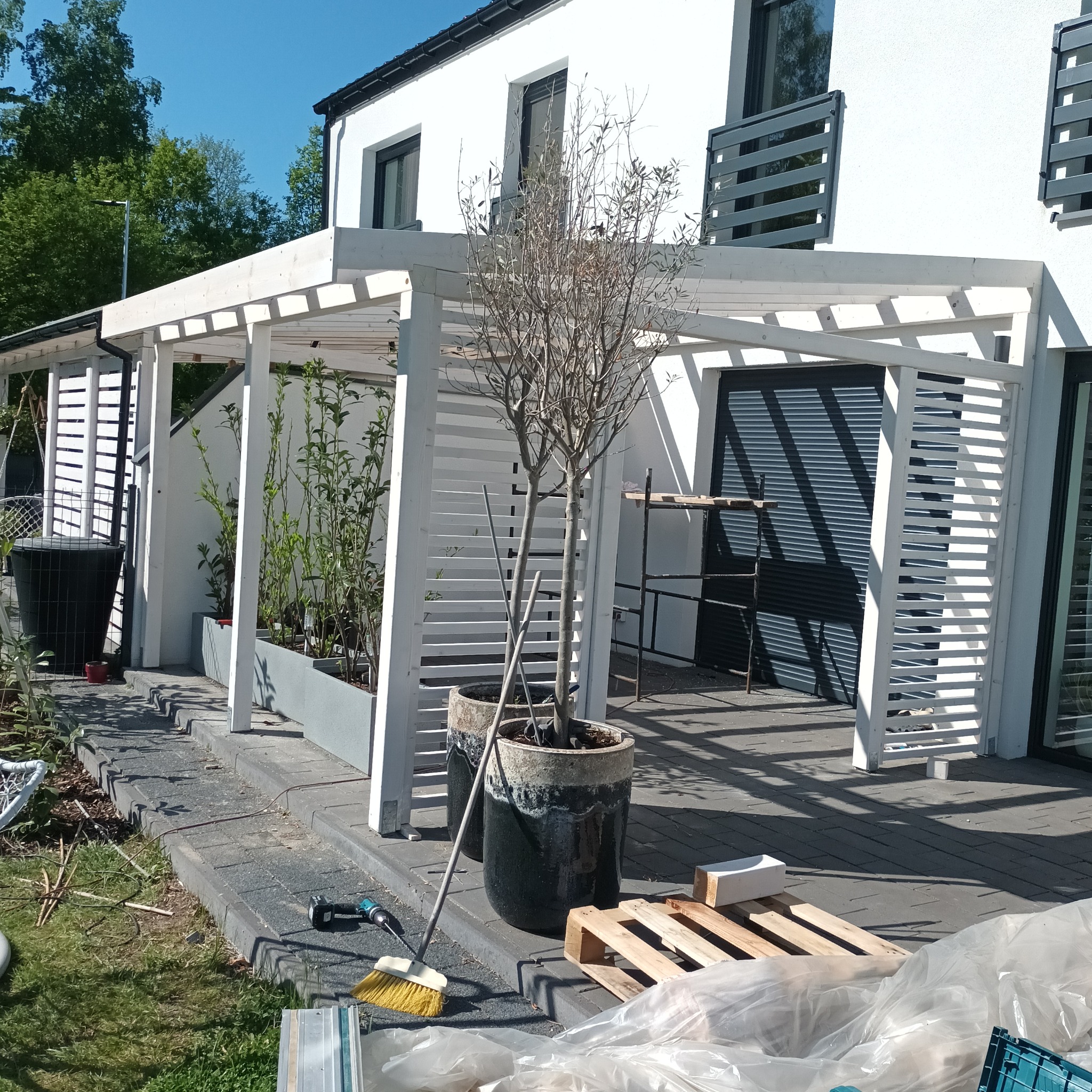 Biała, nowoczesna altana z drewnianymi lamelami przy białym domu. Na pierwszym planie donice z drzewkiem, narzędzia i paleta. Słoneczny dzień.