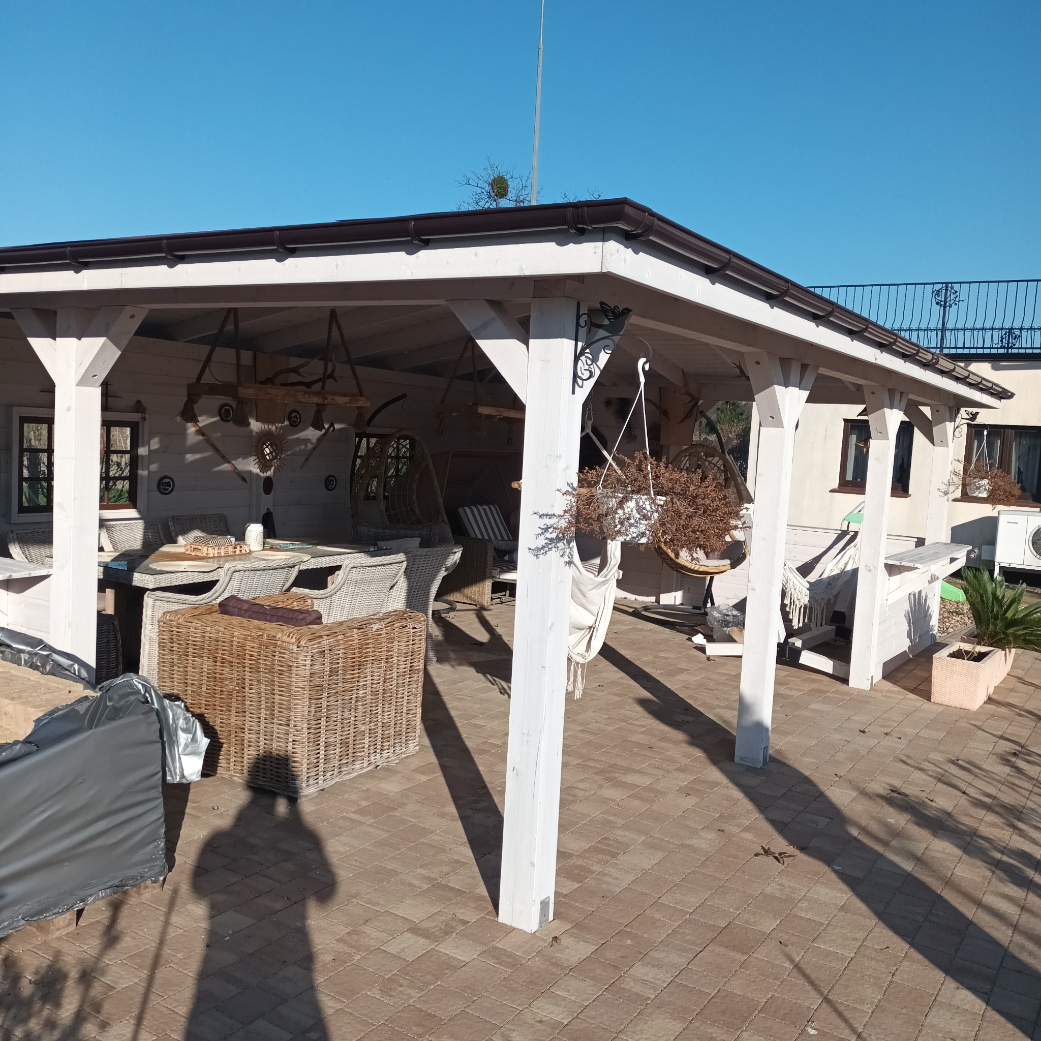 Biała altana z drewnianymi meblami i wiszącymi dekoracjami, oświetlona słońcem. Widoczny cień osoby fotografującej na kostce brukowej.