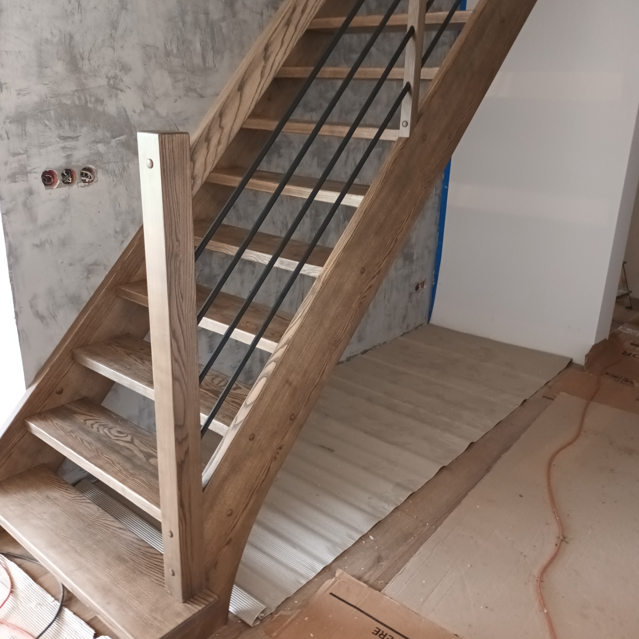Jasne, drewniane schody z czarnymi, metalowymi prętami balustrady, widoczne na tle surowej ściany i przygotowanego podłoża.