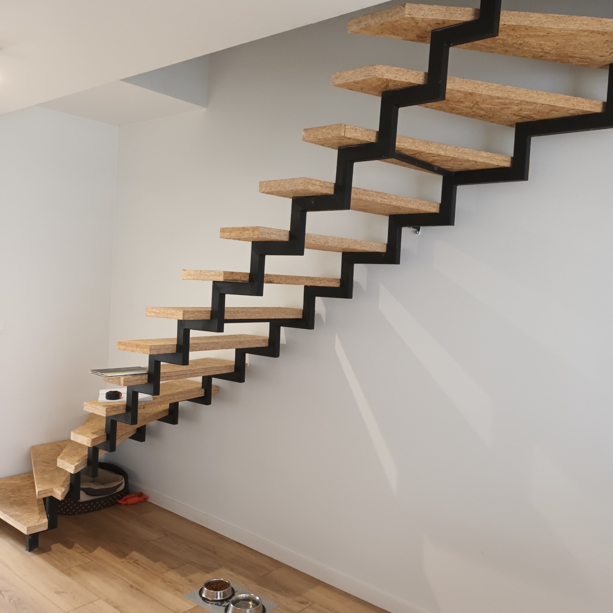 Nowoczesne schody wspornikowe z drewnianymi stopniami i czarną, metalową konstrukcją, widoczne z dołu, z miskami dla zwierząt w dolnym rogu.