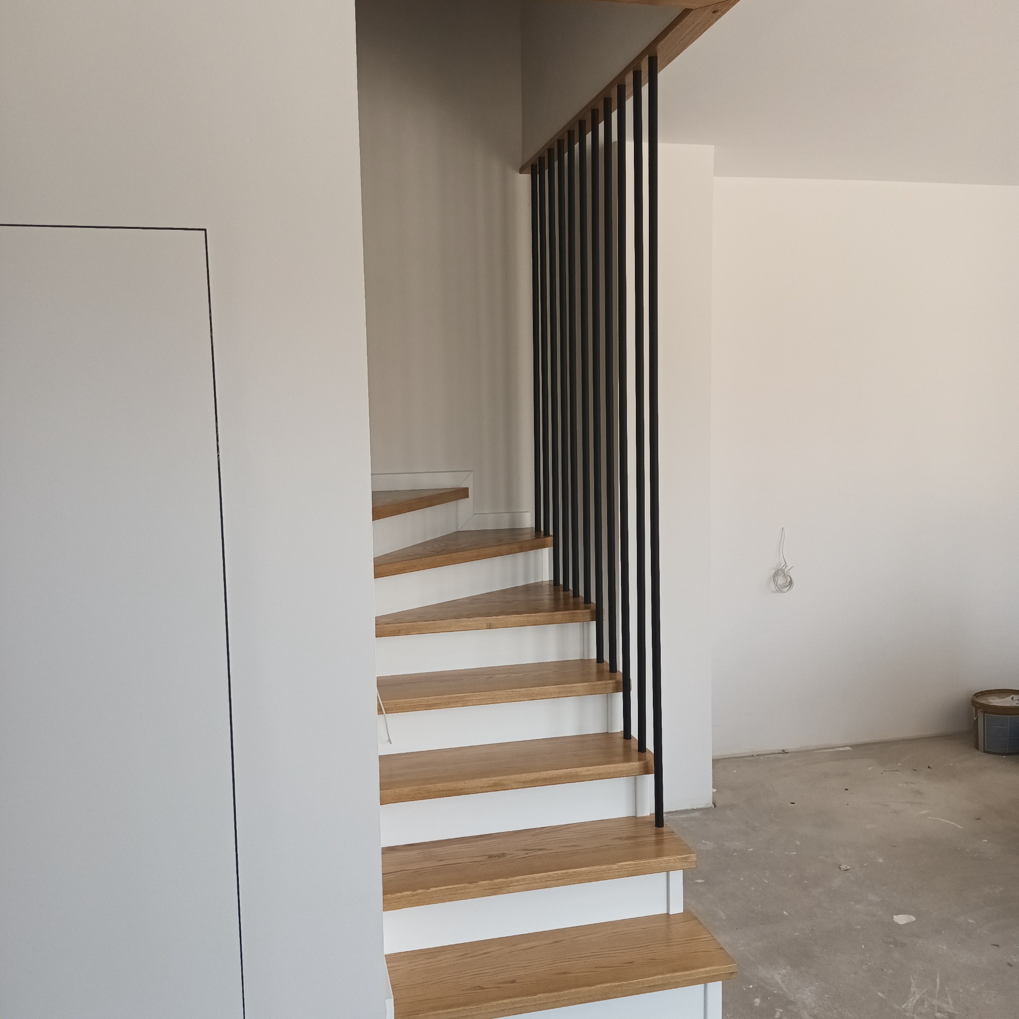 Nowoczesne schody z drewnianymi stopniami i czarnymi, pionowymi poręczami, widok z dołu, surowy beton na podłodze wskazuje na etap wykończeniowy.