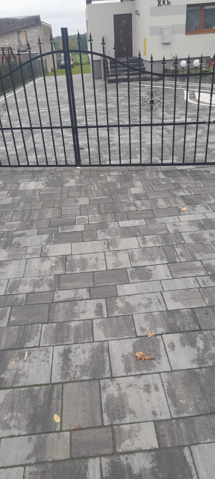 Szara kostka brukowa ułożona w regularny wzór, przed metalową bramą z ozdobnymi elementami, widoczne pojedyncze liście na powierzchni.