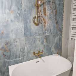 Mianatura sp. Z o.o. - Nowoczesna łazienka z wanną, baterią i deszczownicą w kolorze złotym, ściany wyłożone płytkami imitującymi marmur w odcieniach błękitu i brązu.