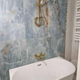 Nowoczesna łazienka z wanną, baterią i deszczownicą w kolorze złotym, ściany wyłożone płytkami imitującymi marmur w odcieniach błękitu i brązu.