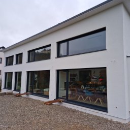 Mianatura sp. Z o.o. - Nowoczesny dom z białym tynkiem strukturalnym i dużymi oknami z czarnymi ramami, widok z zewnątrz na żwirowe podłoże i fragment wnętrza z meblami.