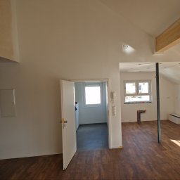 Mianatura sp. Z o.o. - Wnętrze poddasza z drewnianą podłogą, otwartymi drzwiami do jasnego pomieszczenia z oknem roletowym, widoczną instalacją hydrauliczną na ścianie i drewnianą belką stropową.