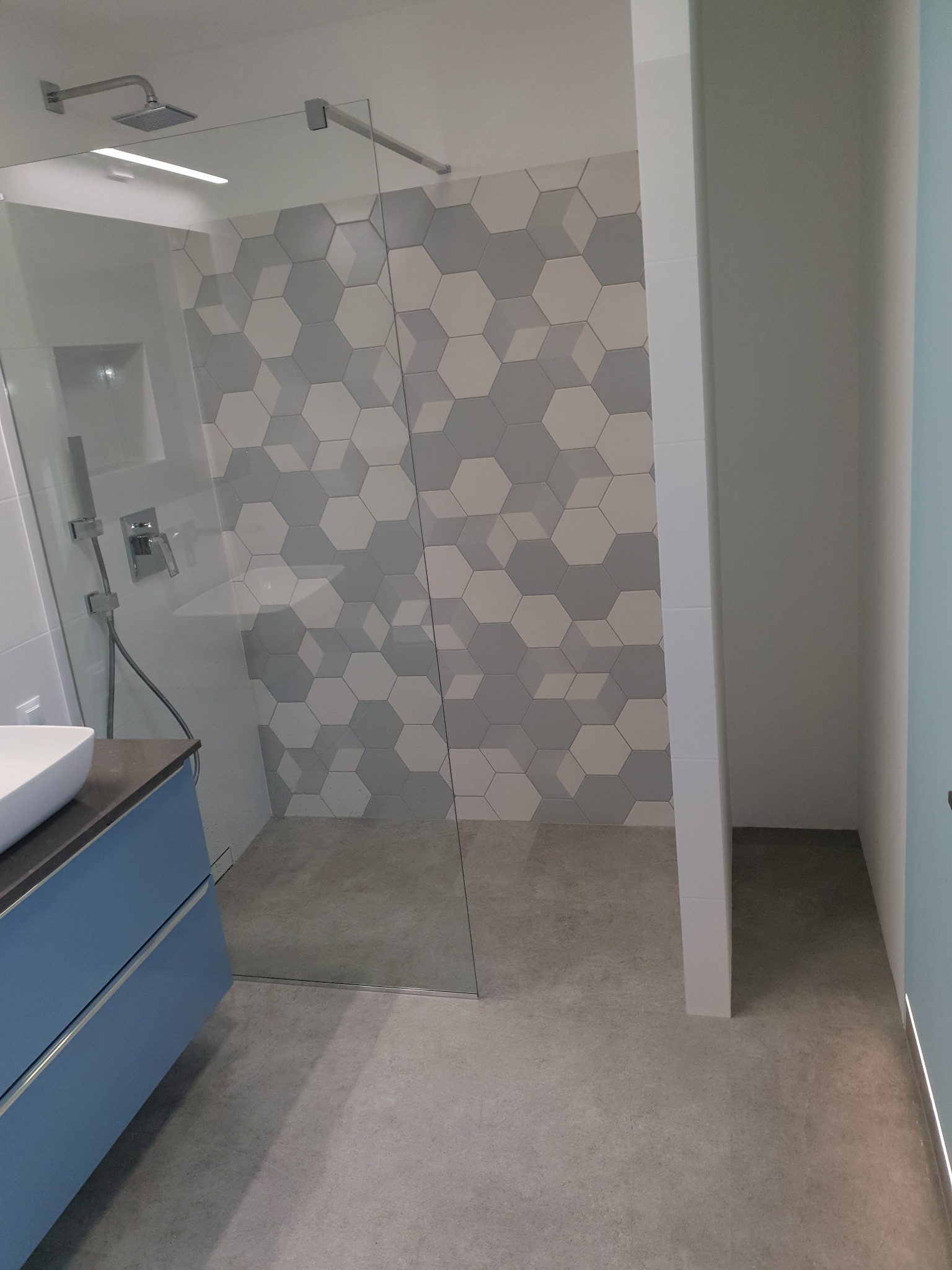 Nowoczesna kabina prysznicowa walk-in z heksagonalnymi płytkami w odcieniach szarości i bieli, umywalka z szafką w kolorze błękitnym, szara podłoga i minimalistyczna armatura.