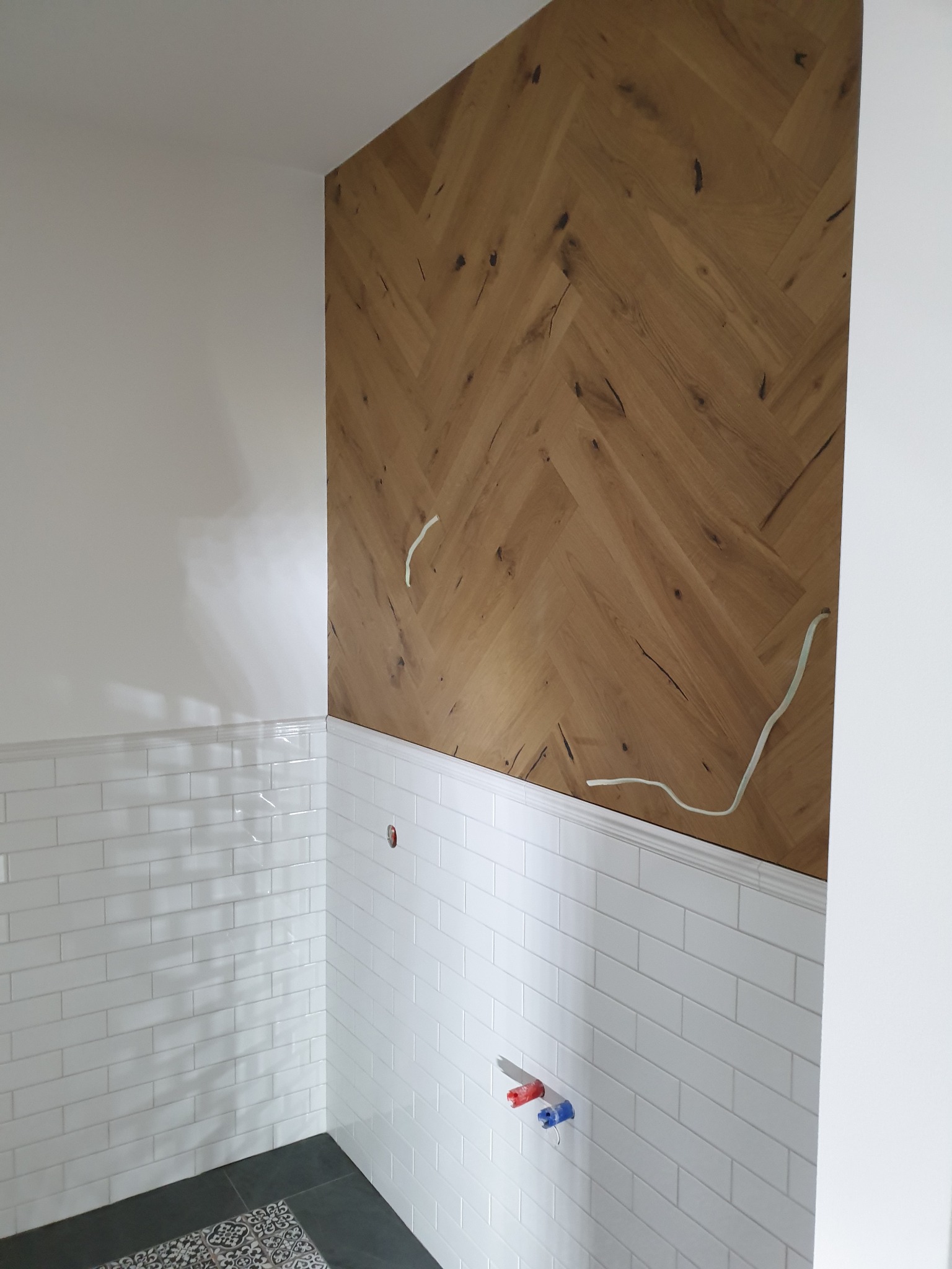 Ściana w łazience wykończona drewnopodobną płytą w jodełkę, białe kafelki na dole ściany z dekoracyjnym paskiem, widoczne wystające kable i instalacja wodna.