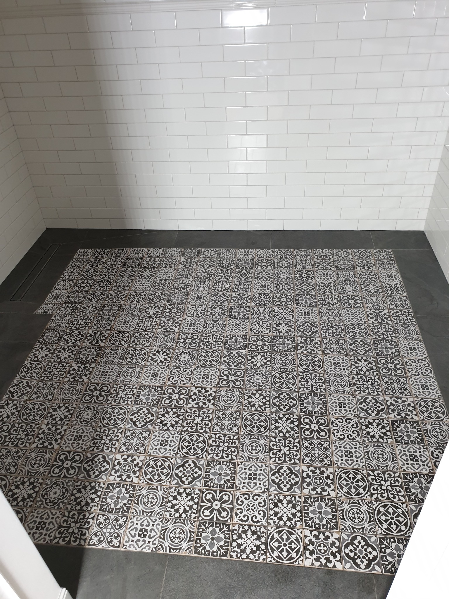 Wykończona podłoga w łazience z czarno-białymi wzorzystymi płytkami, otoczona ciemnoszarą ramką i białymi, prostokątnymi płytkami na ścianach.