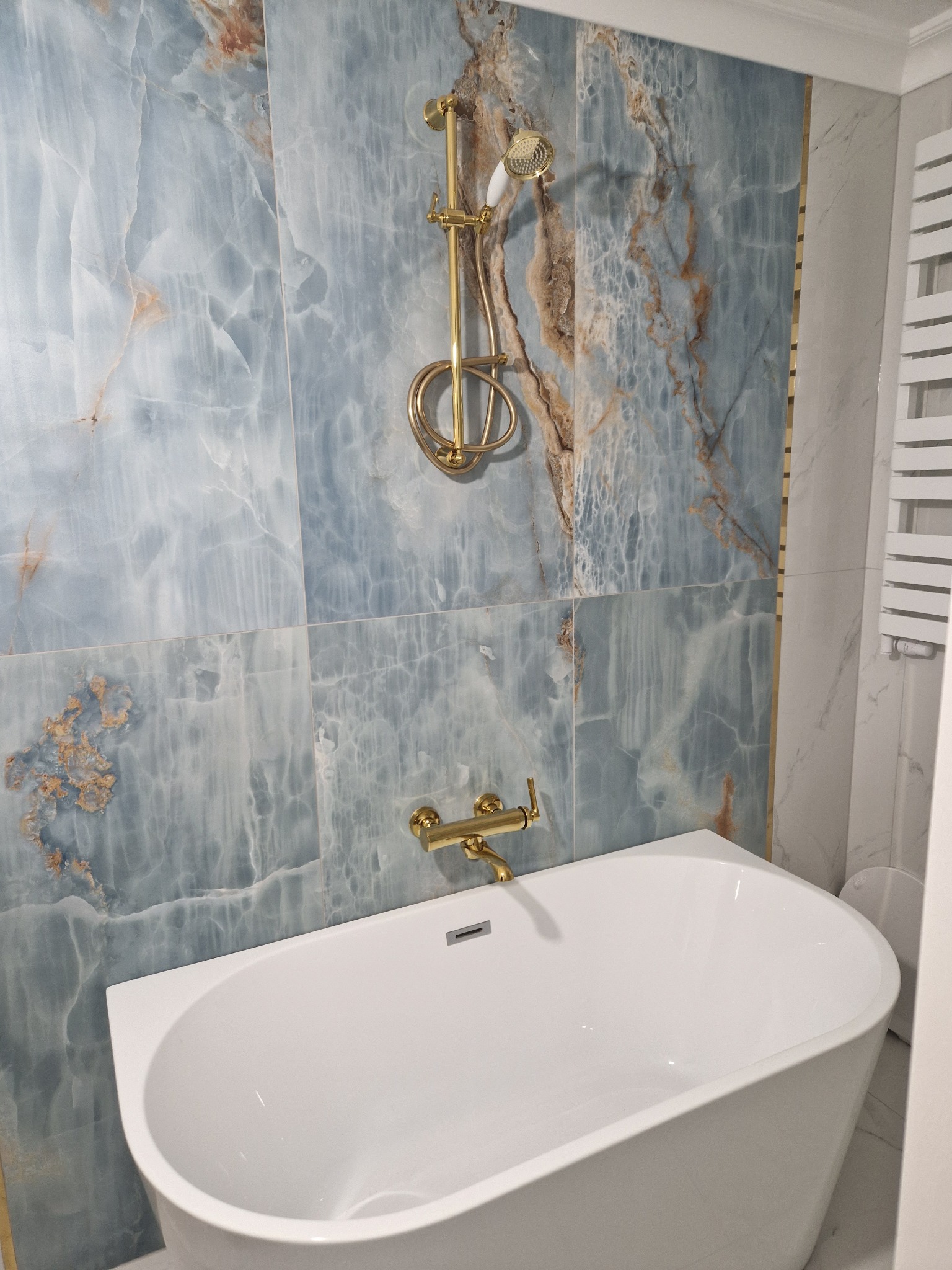 Nowoczesna łazienka z wanną, baterią i deszczownicą w kolorze złotym, ściany wyłożone płytkami imitującymi marmur w odcieniach błękitu i brązu.