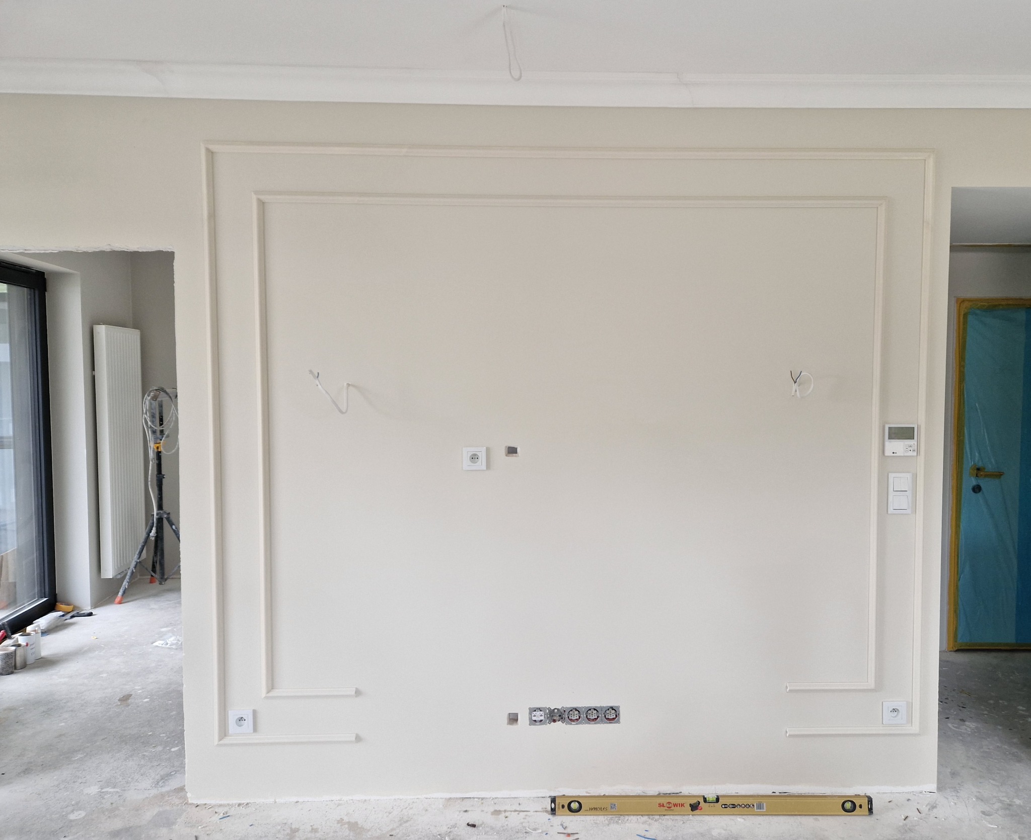 Ściana w trakcie przygotowania do montażu telewizora, z dekoracyjnymi listwami, widoczne gniazdka elektryczne i wyprowadzone kable, poziomica na podłodze, surowy stan wykończenia.