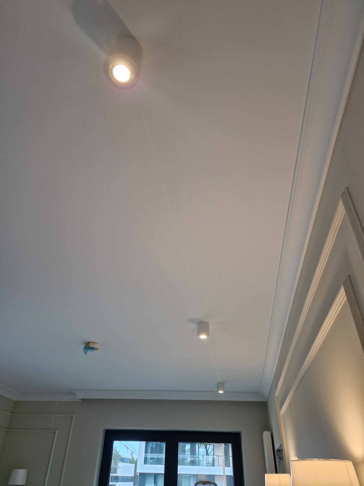 Biały sufit z zamontowanymi trzema cylindrycznymi lampami LED, widoczna listwa dekoracyjna przy ścianie i zabezpieczony czujnik dymu na suficie.