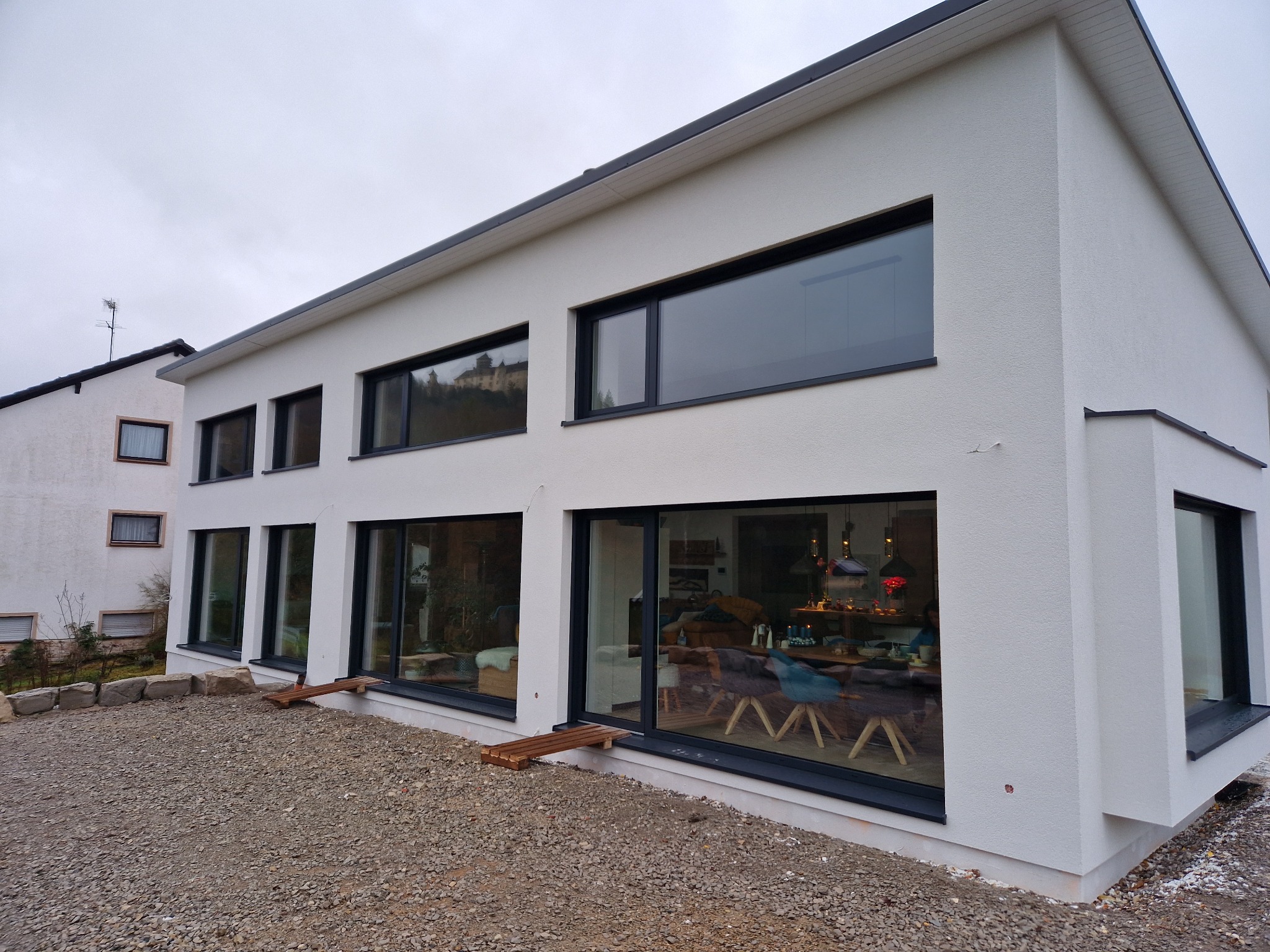 Nowoczesny dom z białym tynkiem strukturalnym i dużymi oknami z czarnymi ramami, widok z zewnątrz na żwirowe podłoże i fragment wnętrza z meblami.