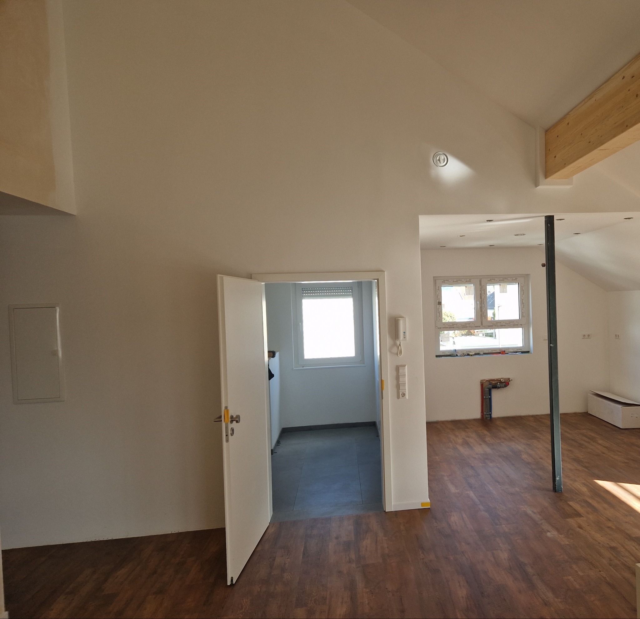 Wnętrze poddasza z drewnianą podłogą, otwartymi drzwiami do jasnego pomieszczenia z oknem roletowym, widoczną instalacją hydrauliczną na ścianie i drewnianą belką stropową.