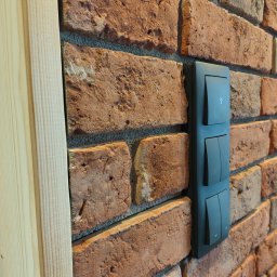 ANTIGUS Adrian KLUCZNY - Czarny, potrójny włącznik światła z ikoną żarówki, zamontowany na dekoracyjnej ścianie z cegły, obok drewnianej listwy wykończeniowej.