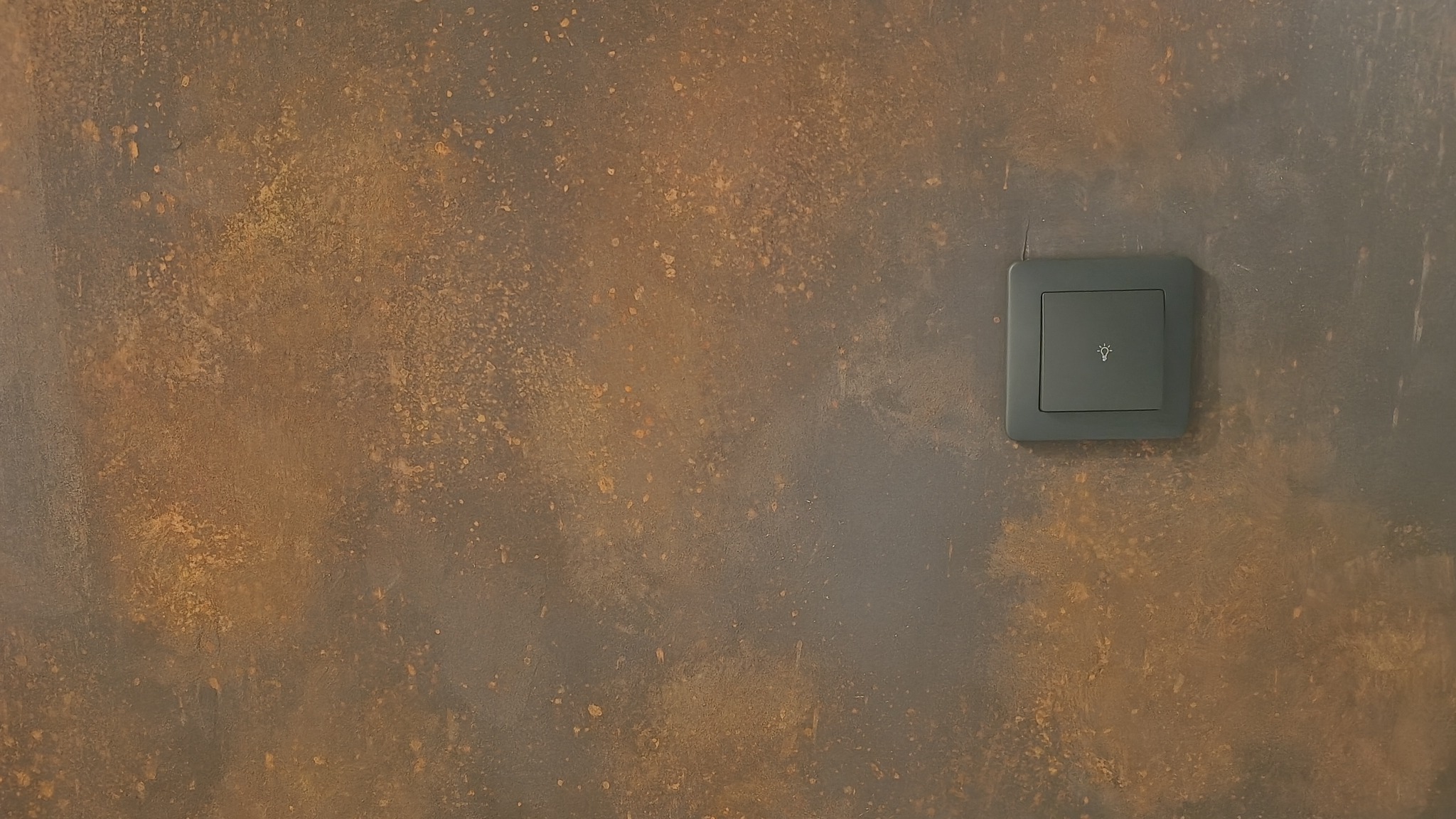 Szary włącznik światła z ikoną żarówki na ścianie o dekoracyjnej, rdzawobrązowej fakturze. Minimalistyczny design w nowoczesnym wnętrzu.