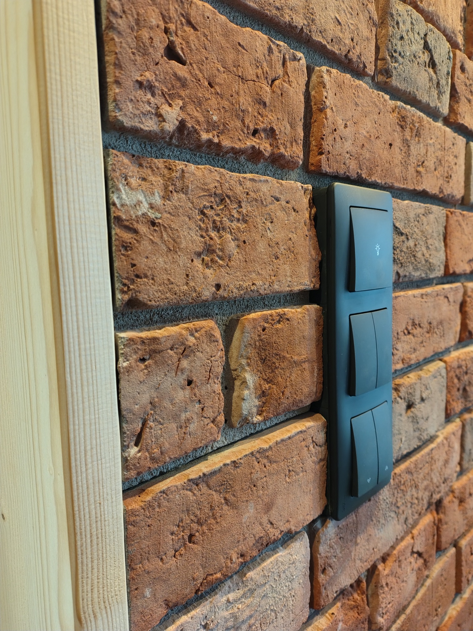 Czarny, potrójny włącznik światła z ikoną żarówki, zamontowany na dekoracyjnej ścianie z cegły, obok drewnianej listwy wykończeniowej.