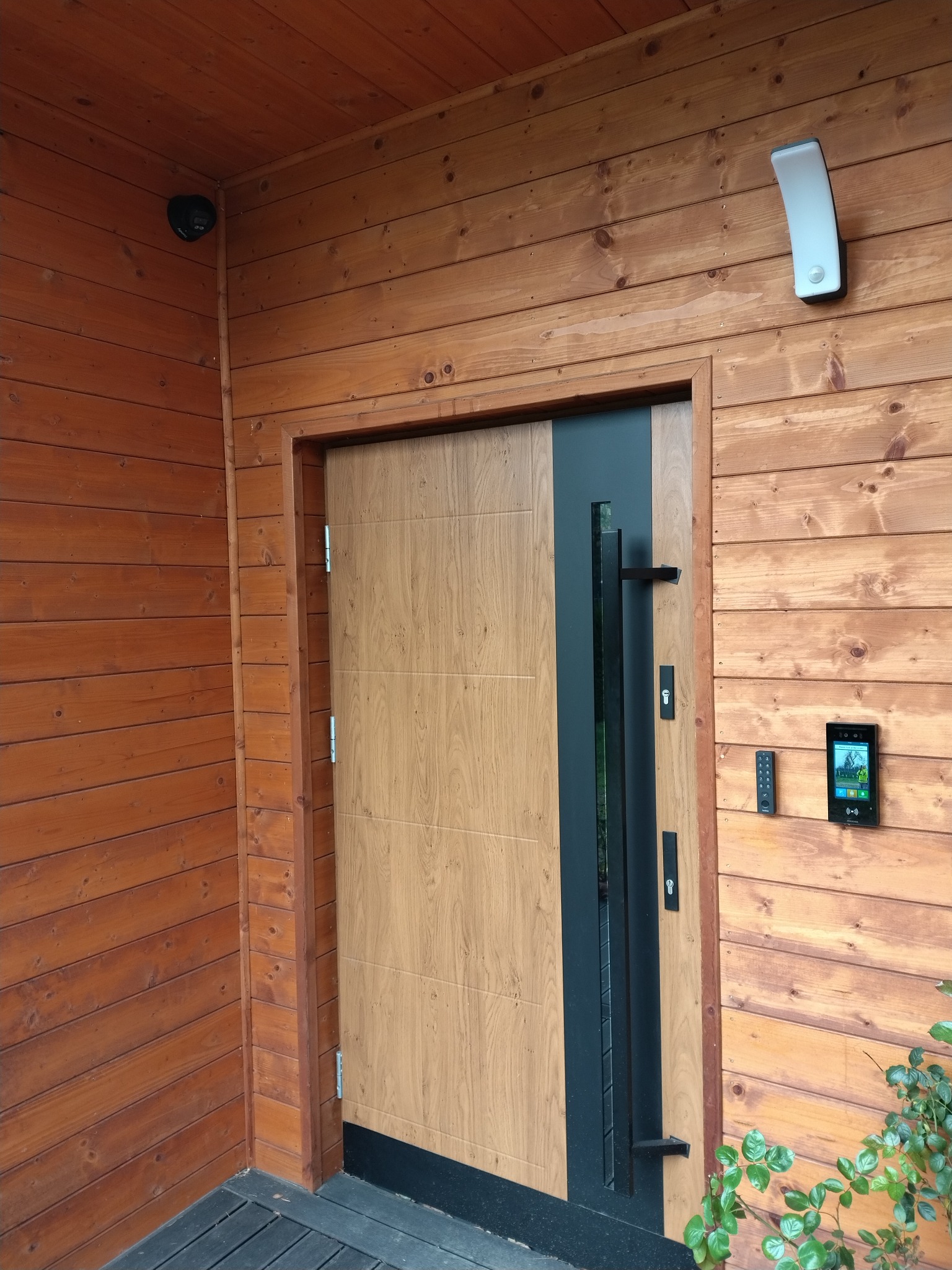 Drewniana elewacja domu z nowoczesnymi drzwiami wejściowymi, wideodomofonem, panelem dotykowym, oświetleniem zewnętrznym i kamerą monitoringu.