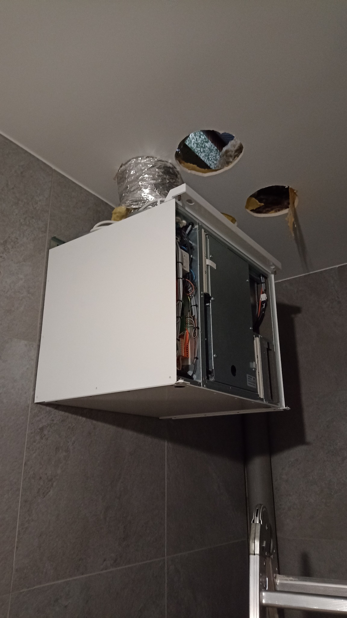 Centrala wentylacyjna z widocznymi przewodami i otworami w suficie, zamontowana w pomieszczeniu z szarą, płytkową ścianą. Widoczny fragment drabiny.