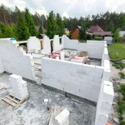 Wznoszenie ścian parteru domu z białych bloczków, widoczny fundament, palety z bloczkami i narzędzia na placu budowy, w tle zieleń drzew i zabudowa jednorodzinna.