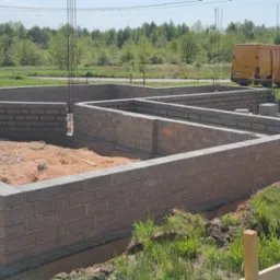 Wzniesione z szarego bloczka fundamenty domu jednorodzinnego, widoczne wystające pręty zbrojeniowe, w tle żółty samochód dostawczy.