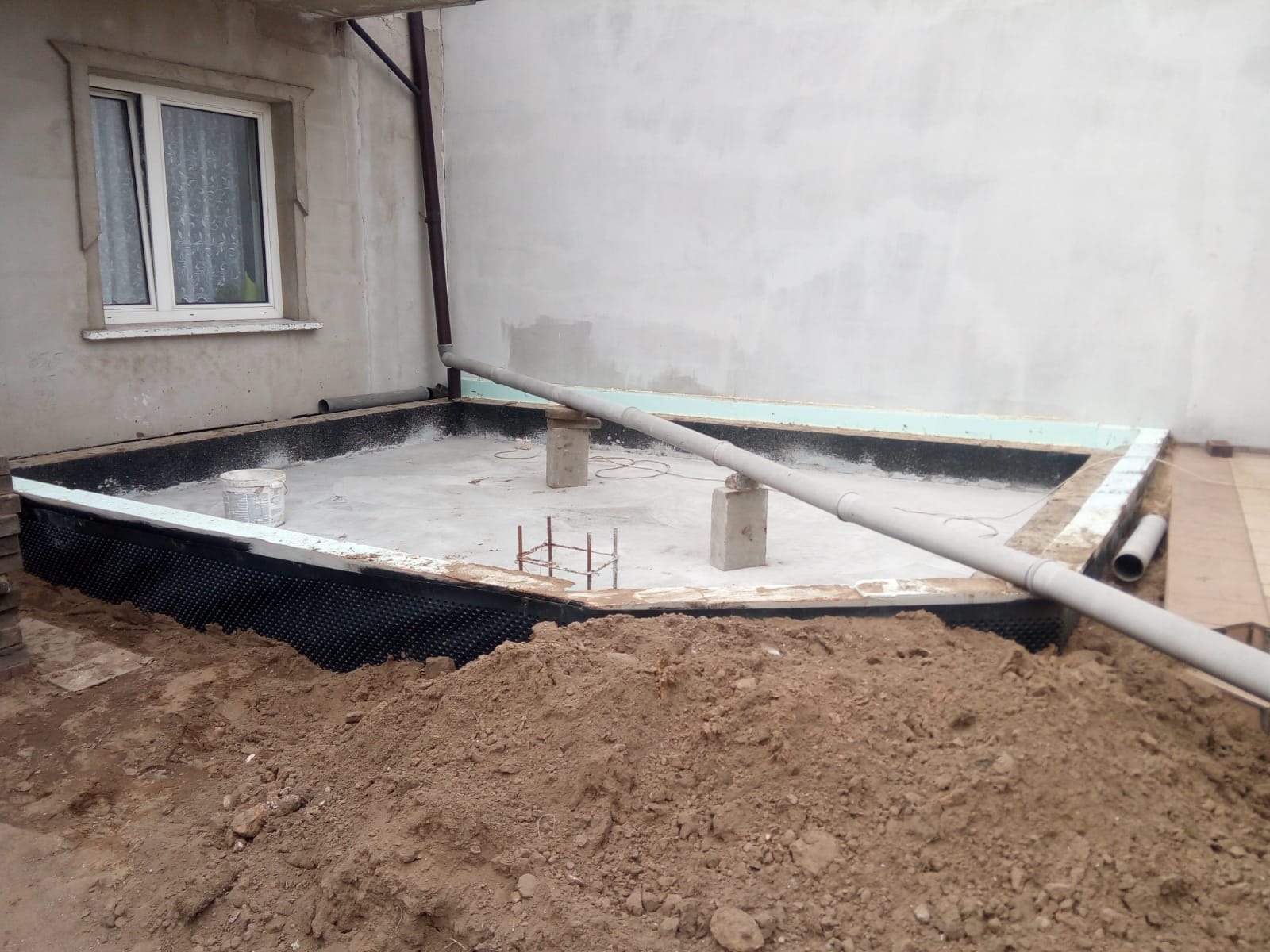Przygotowanie fundamentów: Wykop z izolacją przeciwwilgociową, rury kanalizacyjne, zbrojenie i pryzma ziemi w narożniku budynku.
