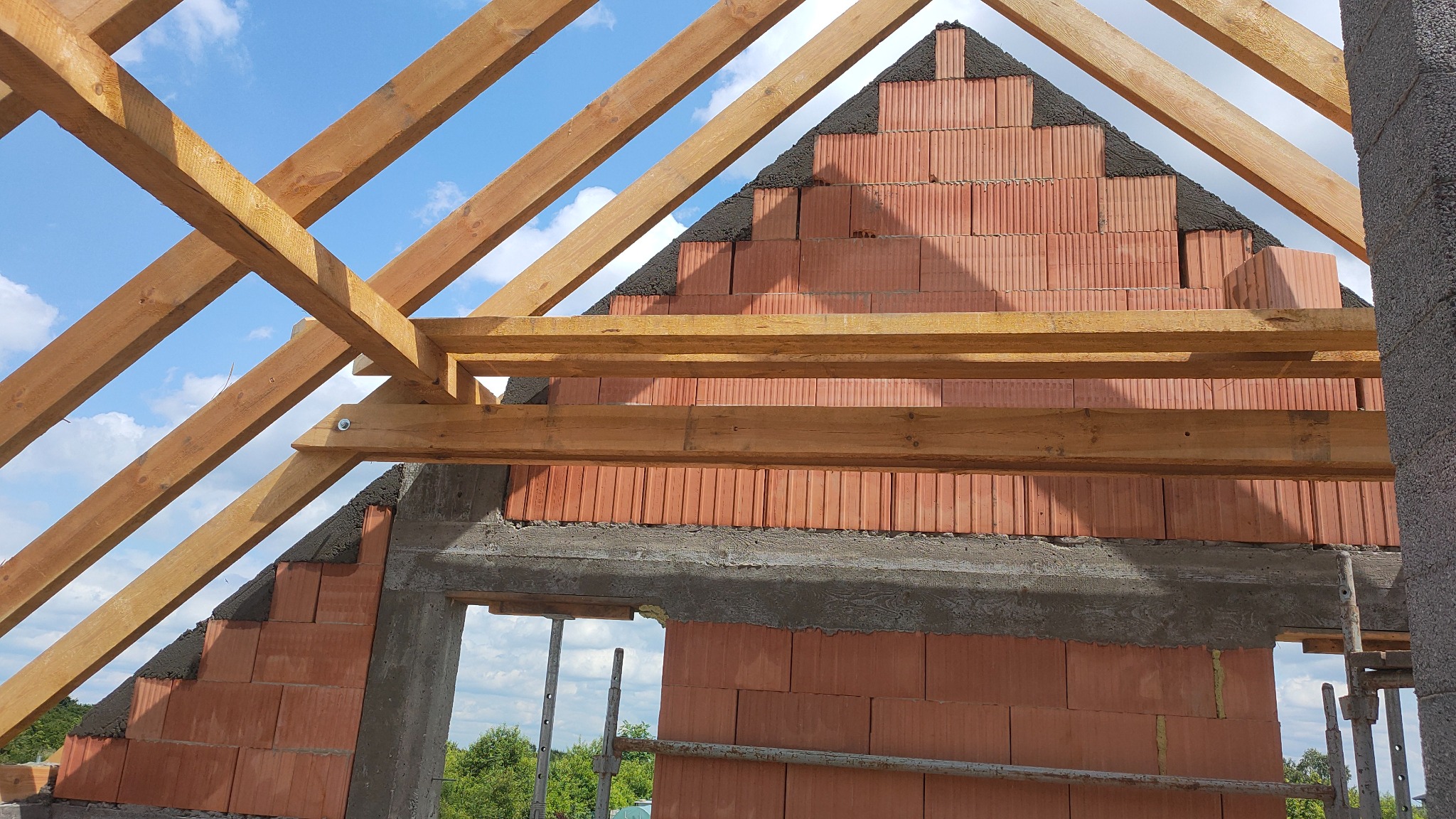 Wznoszenie ścian domu z czerwonej cegły ceramicznej z widoczną konstrukcją drewnianą dachu i fragmentem nieba w tle.