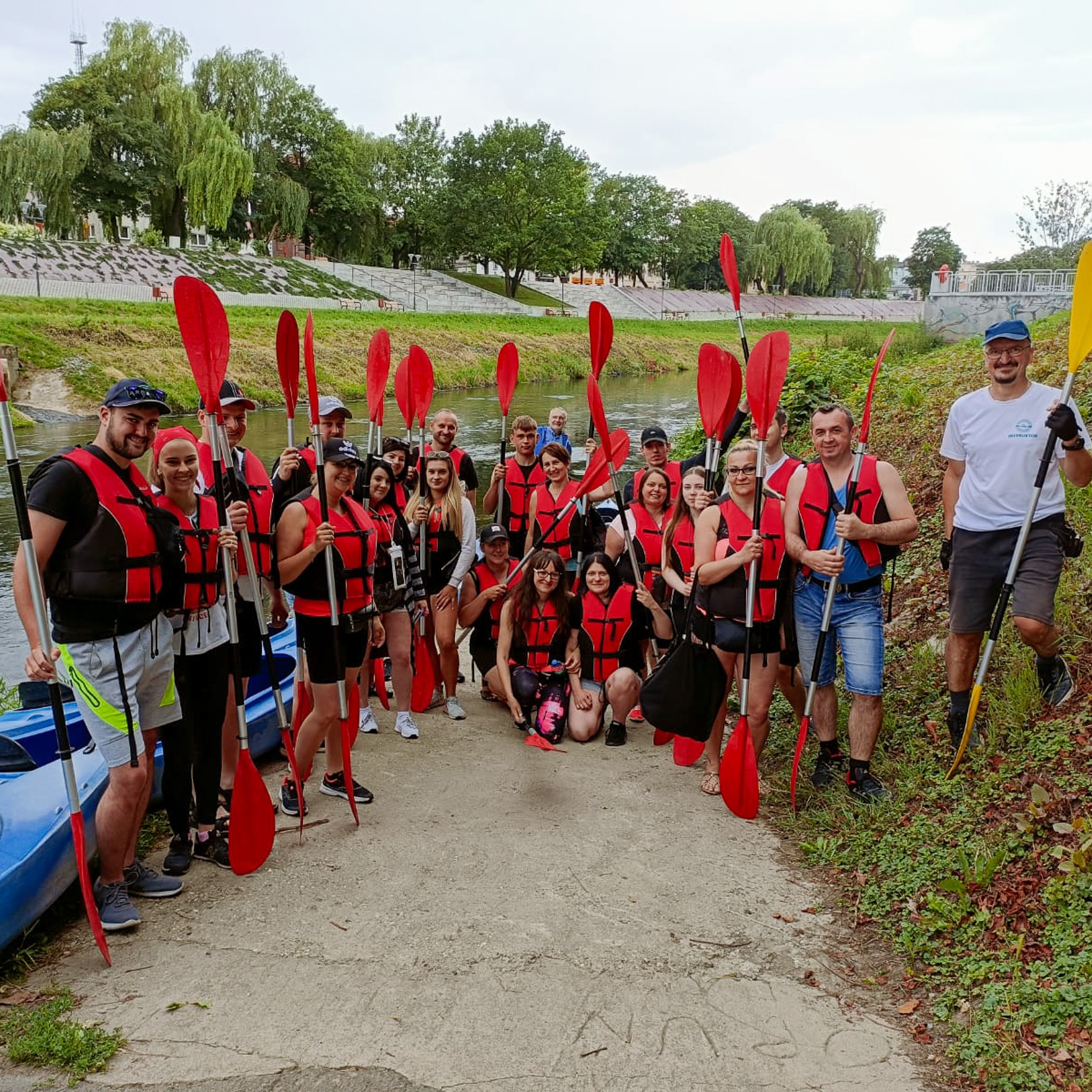 Grupa uśmiechniętych osób w kamizelkach ratunkowych, trzymających czerwone wiosła, gotowa do spływu kajakowego; w tle rzeka i zielony brzeg.