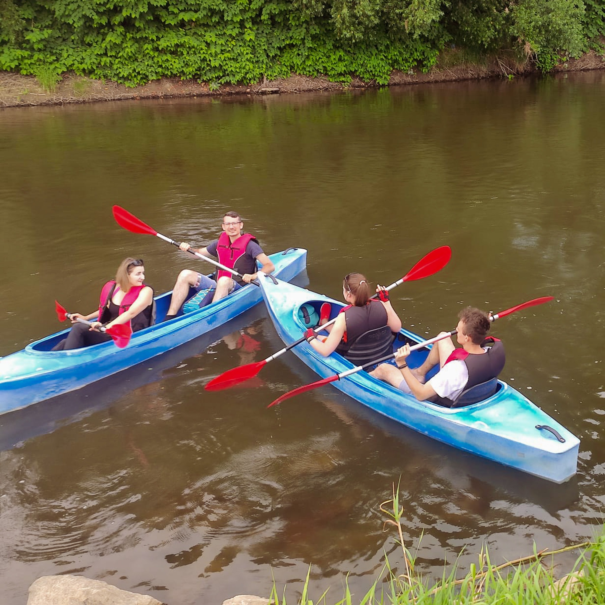 Dwie pary płynące kajakami po rzece w słoneczny dzień, ubrani w kamizelki ratunkowe i wiosłujący czerwonymi wiosłami.