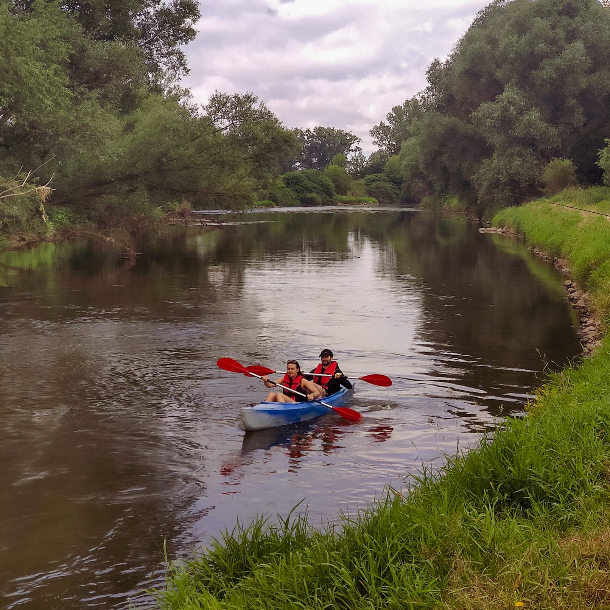 Dwie osoby w niebieskim kajaku płyną po spokojnej rzece otoczonej zielenią, oboje mają na sobie kamizelki ratunkowe i wiosłują czerwonymi wiosłami.
