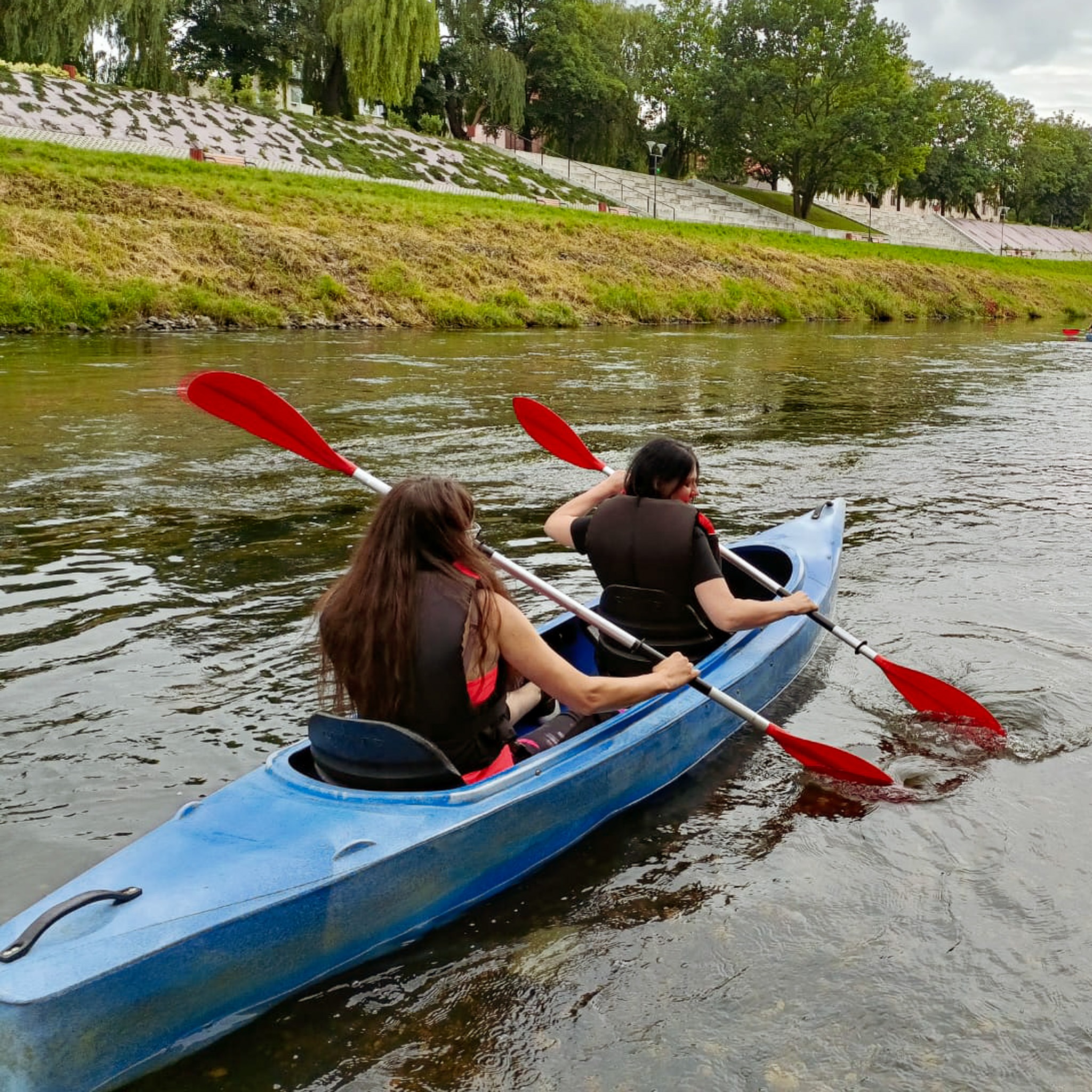 Dwie osoby w kamizelkach ratunkowych płyną kajakiem po rzece w pobliżu zielonego brzegu z drzewami.