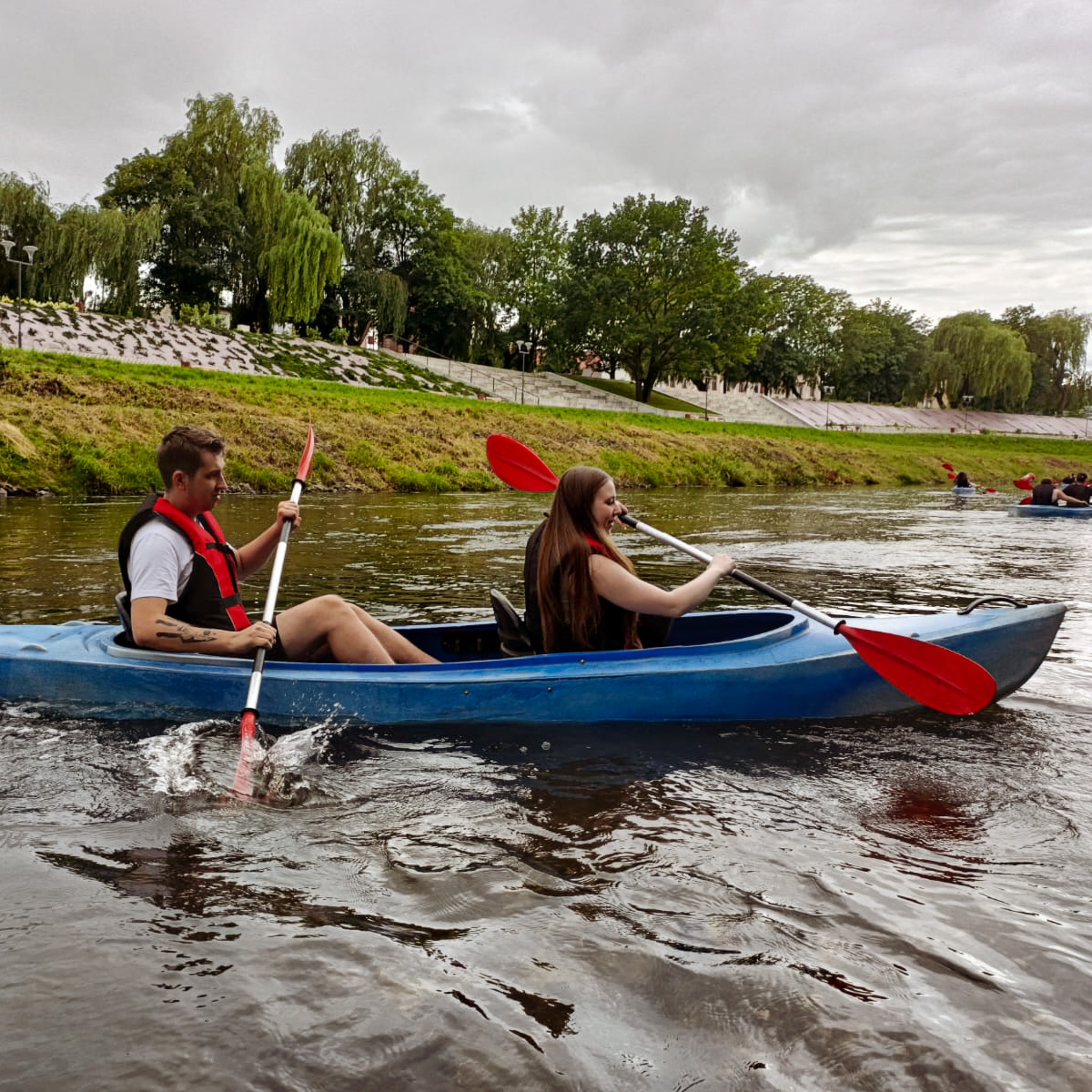 Dwoje kajakarzy w kamizelkach asekuracyjnych wiosłuje w niebieskim kajaku po rzece, w tle zielony brzeg i pochmurne niebo.