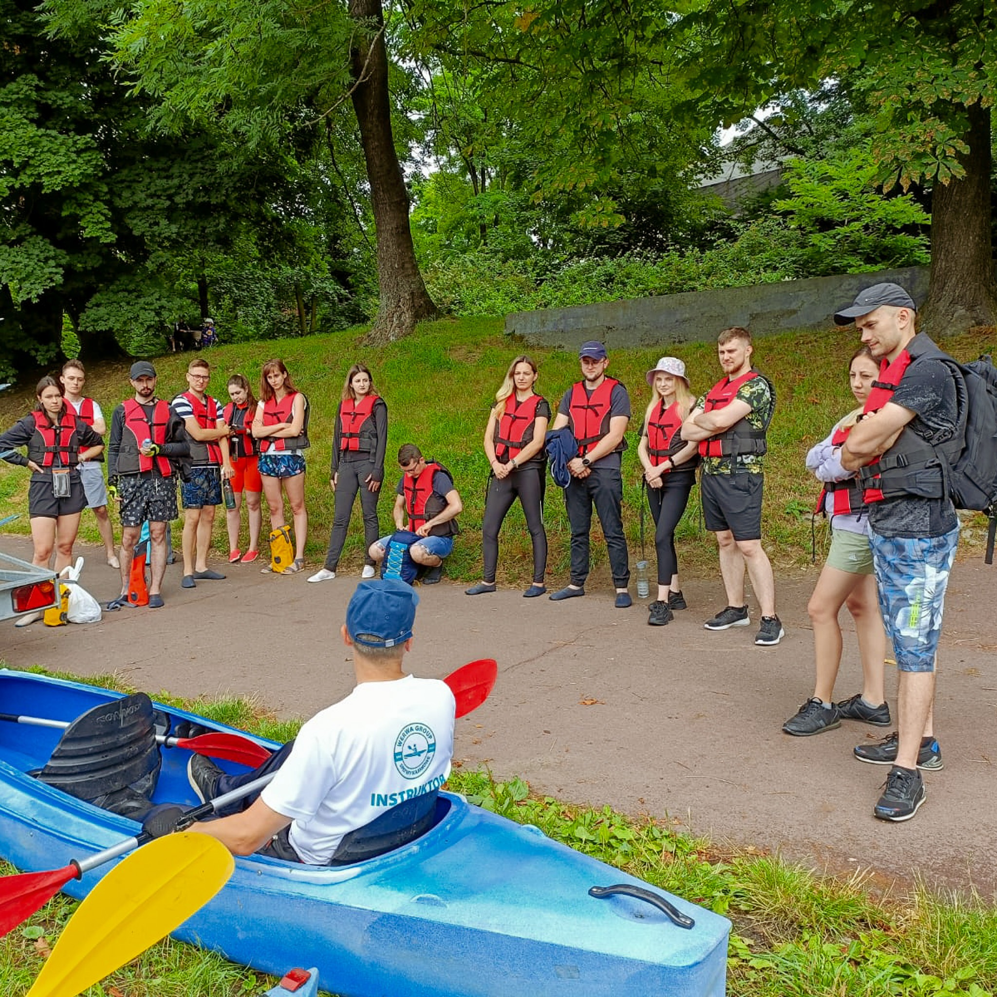 Grupa osób w kamizelkach ratunkowych słucha instruktora siedzącego w niebieskim kajaku z wiosłem na brzegu rzeki, przygotowując się do spływu kajakowego.