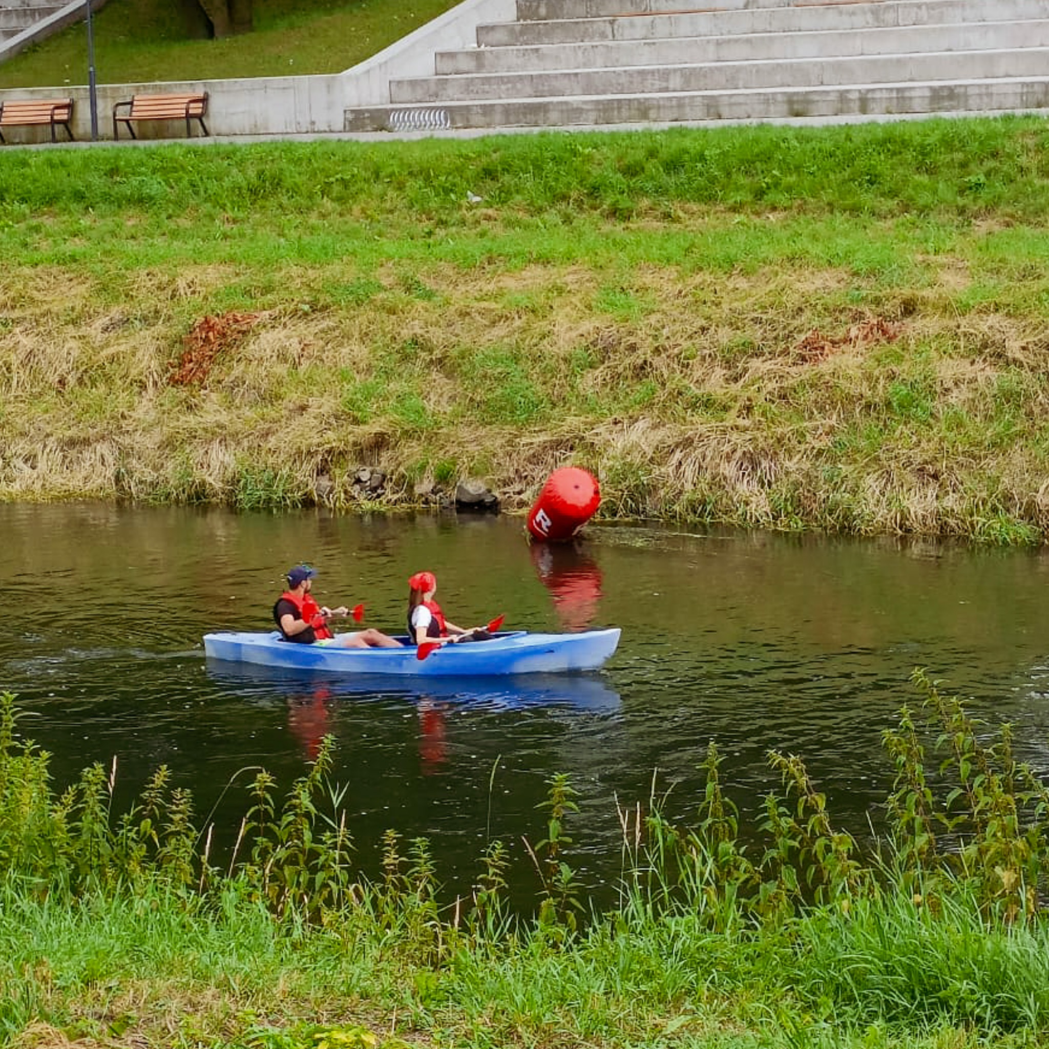 Dwie osoby w czerwonych kamizelkach asekuracyjnych płyną błękitnym kajakiem po spokojnej rzece, na brzegu widoczna bujna roślinność i betonowe schody.