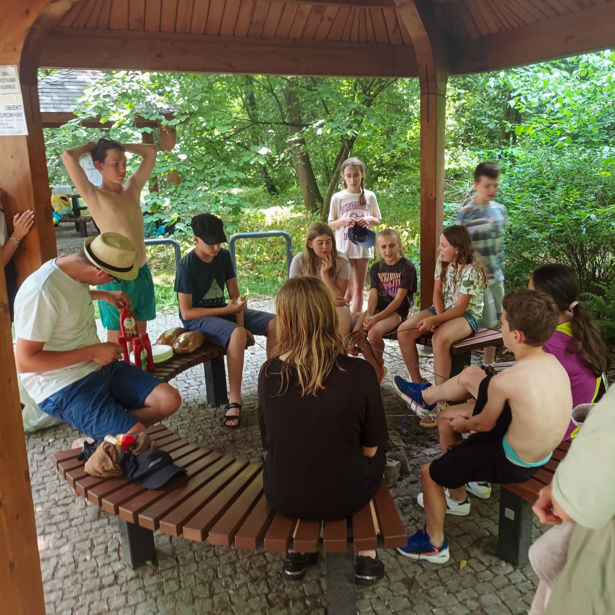 Grupa młodzieży siedząca w altanie, przygotowująca posiłek z ketchupem, w tle zieleń drzew.
