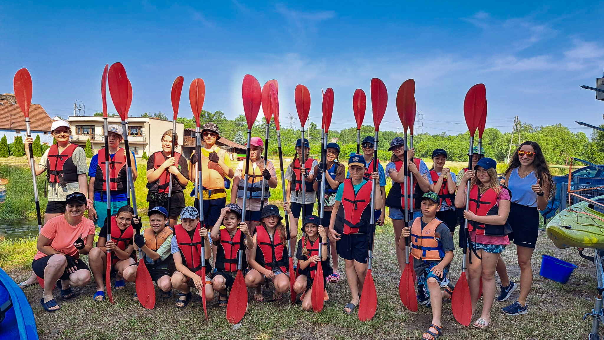Grupa dzieci i dorosłych w kamizelkach ratunkowych trzyma wiosła kajakowe, pozuje do zdjęcia na brzegu rzeki, w tle widoczne budynki i zieleń.