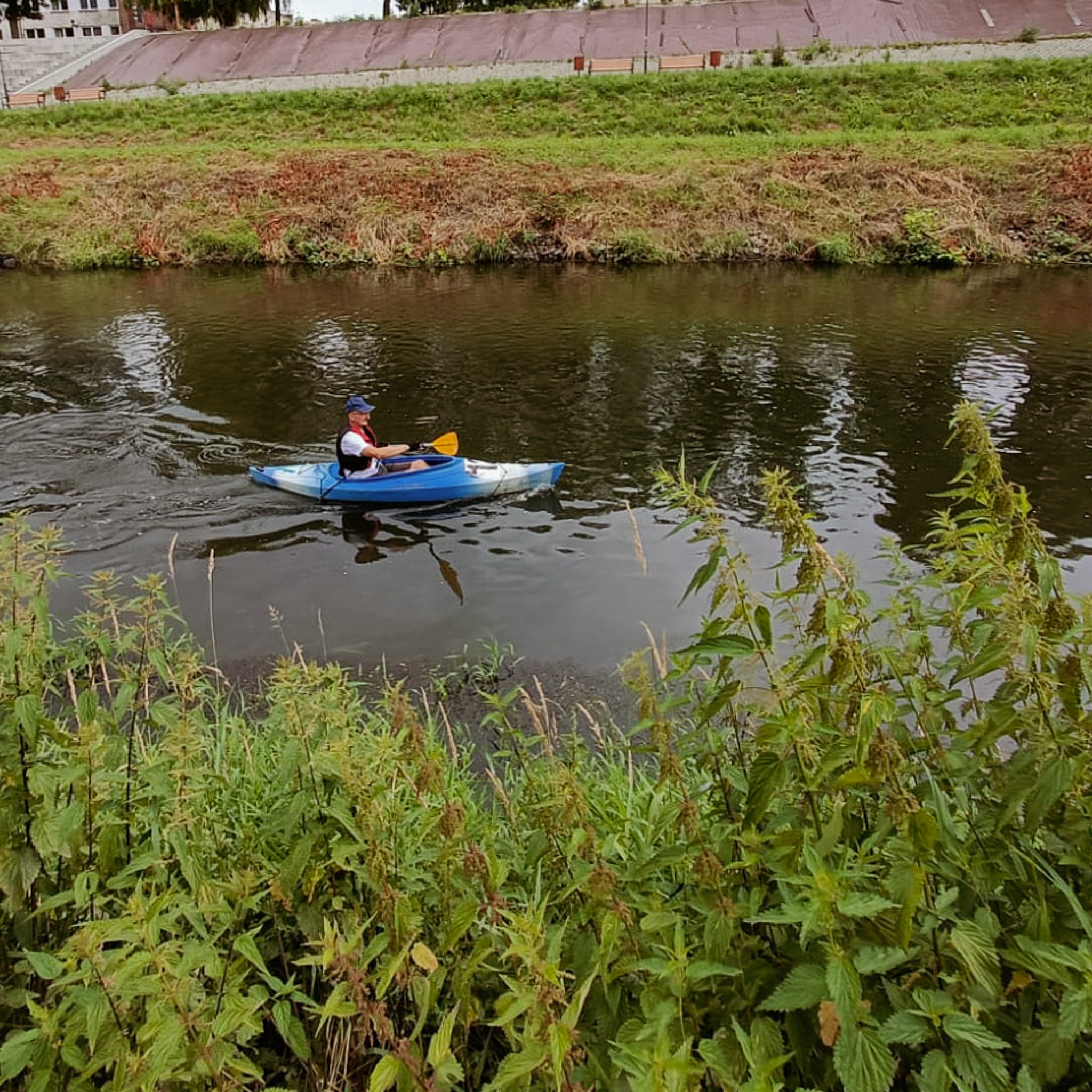 Mężczyzna w kapeluszu płynie kajakiem po spokojnej rzece, widok z brzegu z zaroślami.