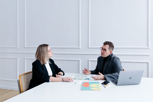 Spotkanie agenta nieruchomości z klientką przy białym stole; omówienie planów mieszkania i dokumentów.