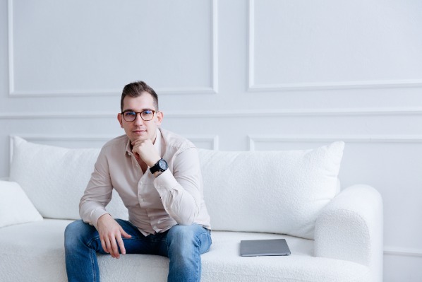 Młody mężczyzna w okularach i koszuli siedzi na białej sofie w minimalistycznym wnętrzu, obok leży laptop.