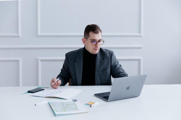 Architekt w okularach analizuje plan mieszkania przy biurku z laptopem, telefonem i długopisami.