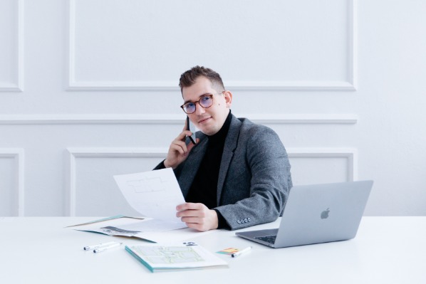 Mężczyzna w okularach i szarej marynarce rozmawia przez telefon trzymając w ręku plan nieruchomości. Na biurku laptop i dokumenty.
