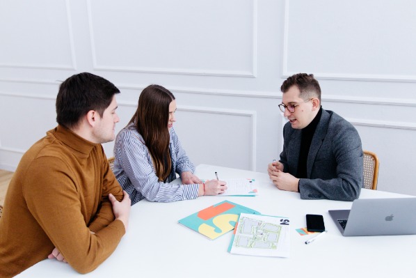 Para podpisuje dokumenty przy białym stole, obok laptop, plan mieszkania i agent nieruchomości w szarym garniturze.