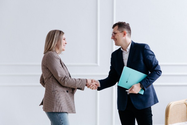 Uśmiechnięci mężczyzna i kobieta w żakietach uściskiem dłoni finalizują umowę, mężczyzna trzyma turkusowy folder z dokumentami na tle białej ściany z dekoracyjnymi panelami.