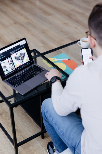 Mężczyzna w okularach siedzi przy szklanym stoliku z laptopem pokazującym galerię zdjęć nieruchomości, obok filiżanka i telefon z klawiaturą numeryczną.