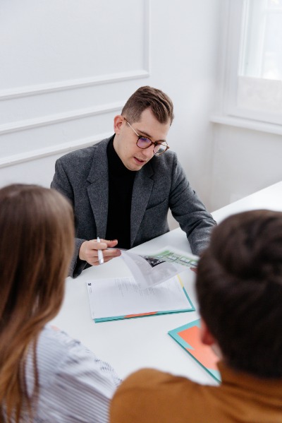 Agent nieruchomości w okularach omawia dokumenty z klientami przy stole, prezentując zdjęcia nieruchomości.