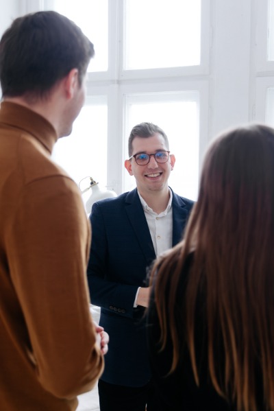 Uśmiechnięty agent nieruchomości w okularach i granatowej marynarce rozmawia z dwoma klientami w jasnym biurze.